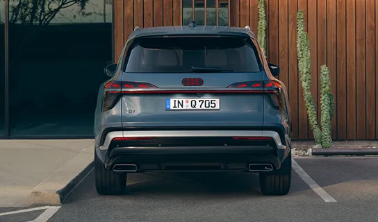 Heckansicht eines blauen Audi Q7 mit durchgehendem Leuchtenband auf einem Parkplatz vor einer Holzfassade.