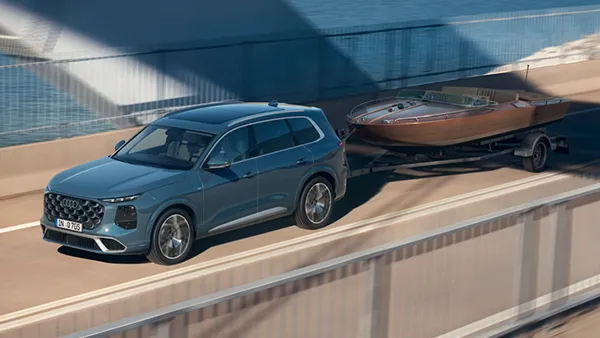 Blauer Audi Q7 zieht auf einer Brücke einen Anhänger mit einem braunen Holzboot.