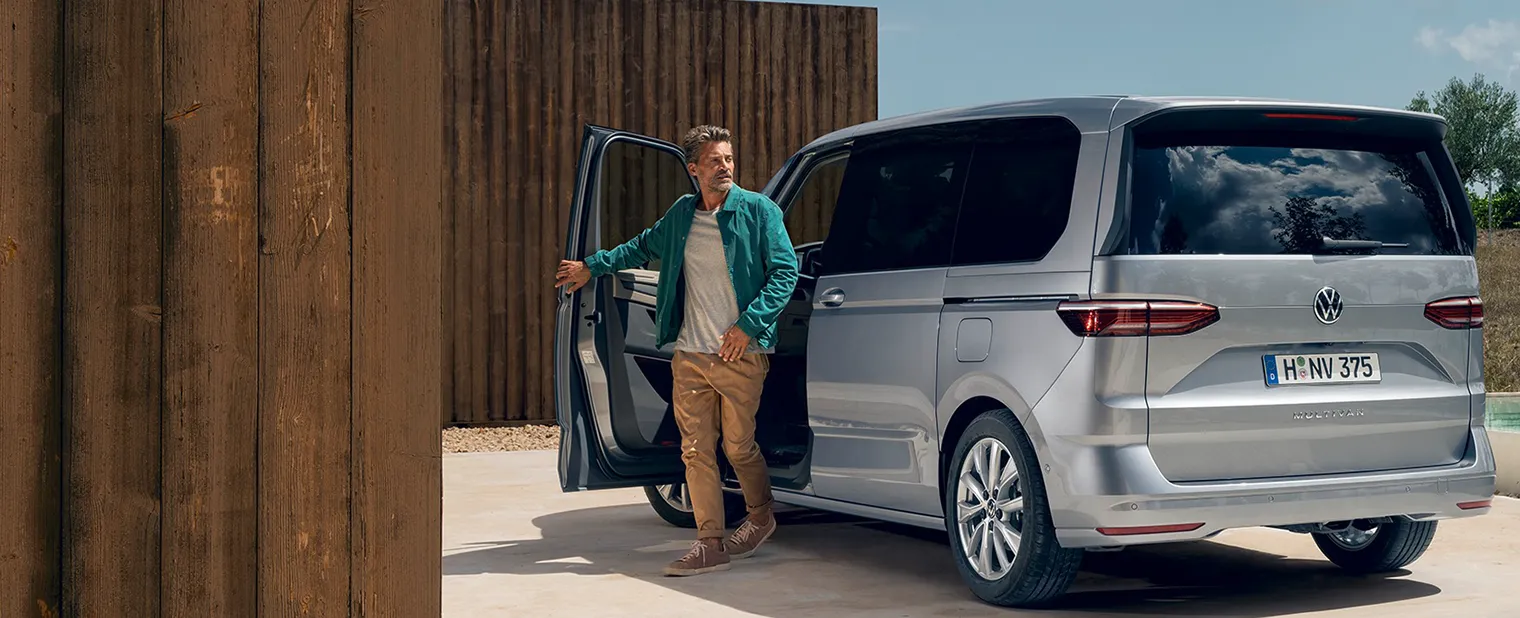 Ein Mann steht neben einem silbernen VW Multivan mit geöffneter Fahrertür vor einer Holzwand.