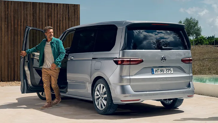 Ein Mann steht neben einem silbernen VW Multivan mit geöffneter Fahrertür vor einer Holzwand.