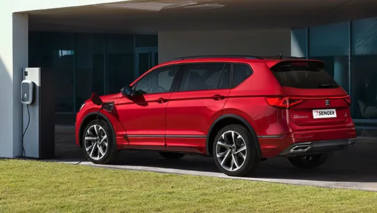 Ein roter SEAT Tarraco e-HYBRID wird an einer Wandladestation vor einem modernen Gebäude geladen.