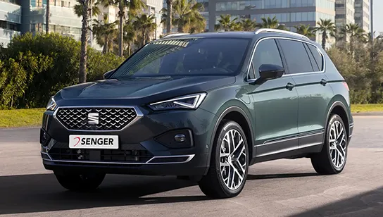 Ein graugrüner SEAT Tarraco steht auf einem Platz vor einer urbanen Kulisse mit Palmen.