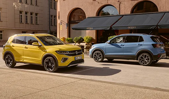 Ein gelber und ein blauer VW T-Cross stehen hintereinander vor einem Gebäude mit Markise und Pflanzen.