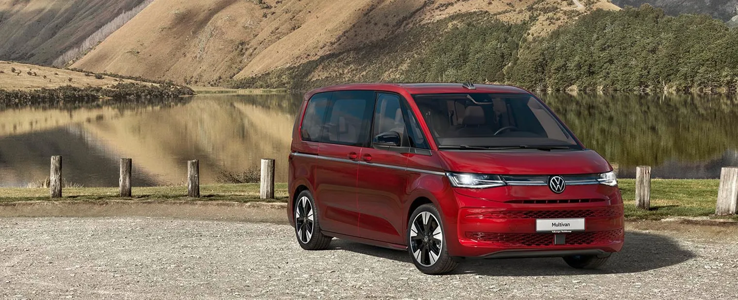 Ein roter VW Multivan steht vor einem See, der von bewaldeten Bergen umgeben ist.