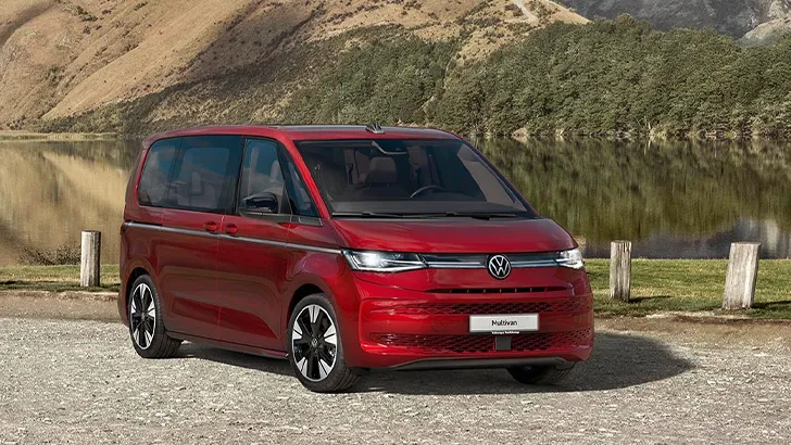 Ein roter VW Multivan steht vor einem See, der von bewaldeten Bergen umgeben ist.