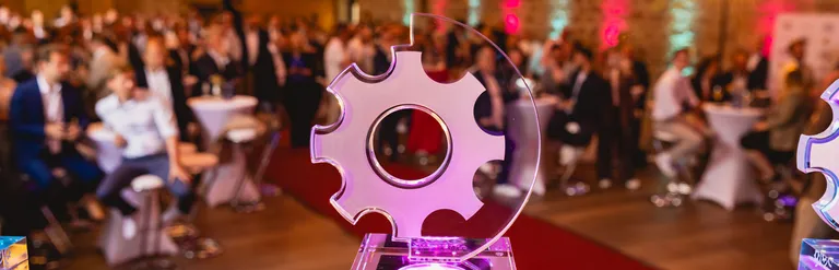 Ein Award in Form eines Zahnrades steht auf einer Bühne, im Hintergrund sitzen viele Menschen an Tischen.