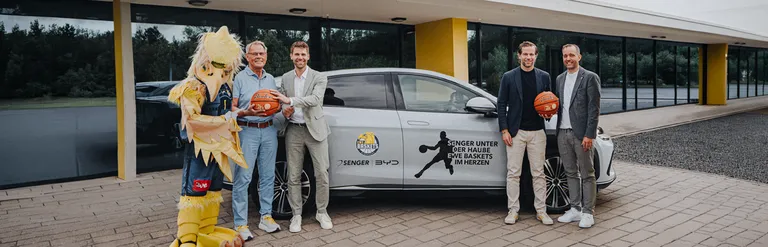Vier Männer in Anzügen und ein Maskottchen im gelben Vogelkostüm mit Basketball von Senger und den EWE Baskets posieren vor einem silbernen BYD-Modell.