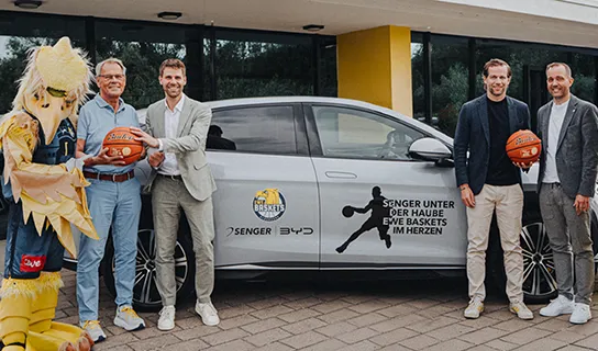 Vier Männer in Anzügen und ein Maskottchen im gelben Vogelkostüm mit Basketball von Senger und den EWE Baskets posieren vor einem silbernen BYD-Modell.