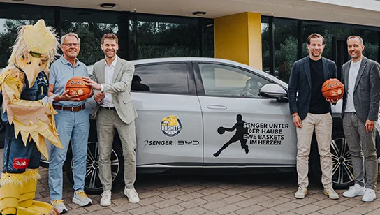Vier Männer in Anzügen und ein Maskottchen im gelben Vogelkostüm mit Basketball von Senger und den EWE Baskets posieren vor einem silbernen BYD-Modell.