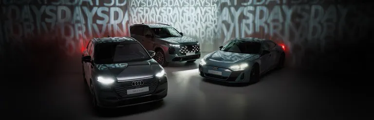 Der Audi Q4 e-tron, Audi e-tron GT und Audi Q7 stehen mit eingeschalteten Scheinwerfern vor einer Wand mit wiederholtem Schriftzug 'DRIVING DAYS'.