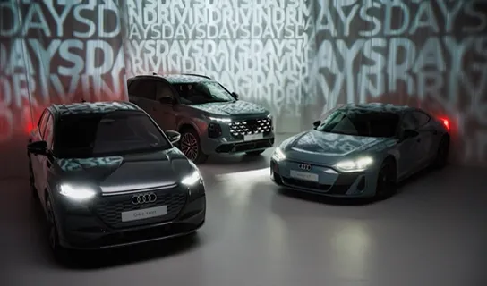 Der Audi Q4 e-tron, Audi e-tron GT und Audi Q7 stehen mit eingeschalteten Scheinwerfern vor einer Wand mit wiederholtem Schriftzug 'DRIVING DAYS'.