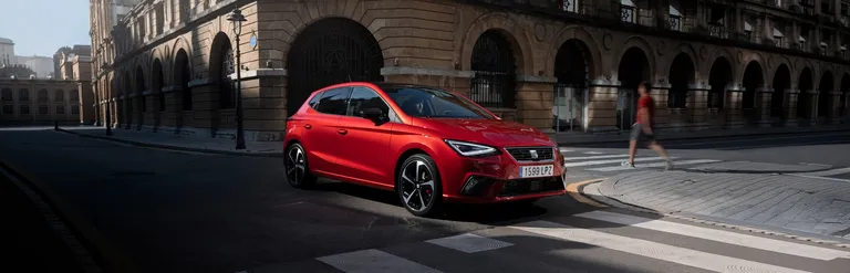 Ein roter SEAT Ibiza steht vor einem Zebrastreifen vor einem historischen Gebäude, daneben geht eine Personen über einen weiteren Zebrastreifen.