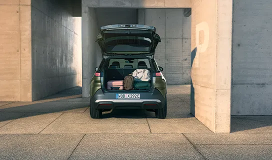Ein VW ID. Cross in grün-metallic steht mit geöffnetem Kofferraum und mehreren Koffern darin vor einem modernen, hellen Betonbau.