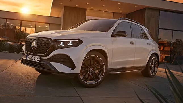 Ein weißer Mercedes GLE SUV steht vor einem modernem Gebäude mit Glasfassade und flachem Dach bei Sonnenuntergang.