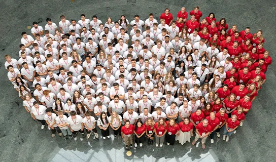Eine große Gruppe von Menschen steht auf einem grauen Platz. Die größere Gruppe links, bei der alle weiße T-Shirts tragen, bildet einen Kreis und die kleinere Gruppe rechts, bei der alle rote T-Shirts tragen, steht in Form eines Bogens.
