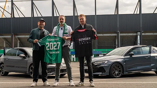 Drei Männer stehen vor zwei grauen Autos mit Senger-Logo, im Hintergrund ist ein Stadion zu sehen. Der linke und der mittlere Mann halte ein grünes SC Preußen Münster-Trikot mit der Aufschrift 'Münster 2027 Senger', der rechte ein schwarzes Senger-Trikot.