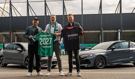 Drei Männer stehen vor zwei grauen Autos mit Senger-Logo, im Hintergrund ist ein Stadion zu sehen. Der linke und der mittlere Mann halte ein grünes SC Preußen Münster-Trikot mit der Aufschrift 'Münster 2027 Senger', der rechte ein schwarzes Senger-Trikot.