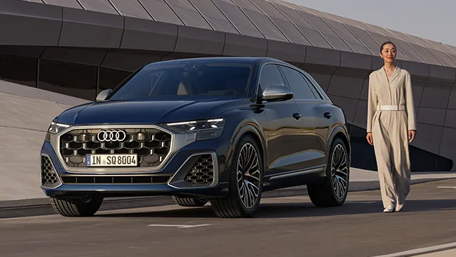 Seitliche Ansicht eines blauen Audi SQ8 auf einer Straße vor einer modernen Architektur, daneben eine stehende Frau in hellem Anzug.