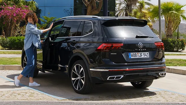 Ein schwarzer VW Tiguan Allspace steht auf einem Parkplatz vor einem modernen Gebäude, eine Frau öffnet die Fahrertür.
