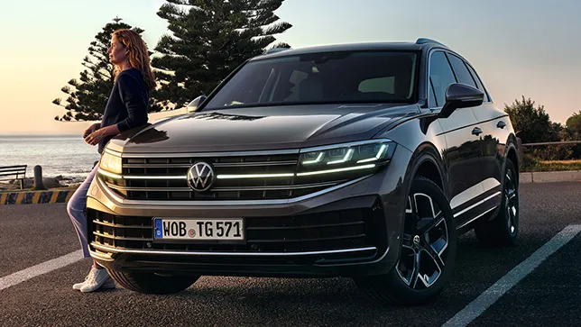 Frontansicht eines grauen VW Touareg auf einem Parkplatz stehend. Ein Frau steht an der linken Seite.