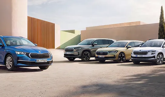 Vier verschiedene Škoda-Modelle in Blau, Grau, Gold und Weiß stehen nebeneinander vor moderner Architektur mit klaren Linien und Bäumen im Hintergrund.